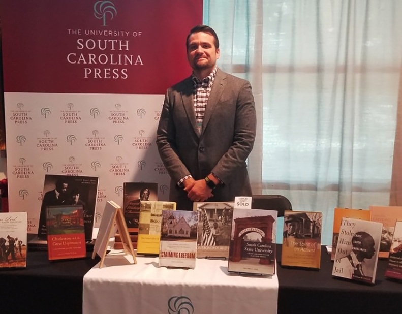 Man wearing suit stands behind table with books on stands. A logo pop-up banner stands behind the table and man.